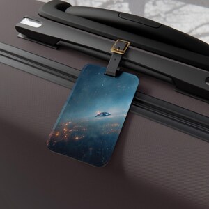May include: A rectangular luggage tag featuring a space scene with a flying saucer over a city at night. The tag is attached to a black handle with a gold-colored buckle. The background is a dark gray suitcase.