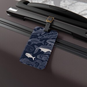 Może przedstawiać: Granatowa etykieta bagażowa z biało-niebieską ilustracją różnych stworzeń morskich. Etykieta jest prostokątna z zaokrąglonymi rogami i czarnym paskiem ze złotą klamrą. Projekt zawiera wieloryby i ryby.