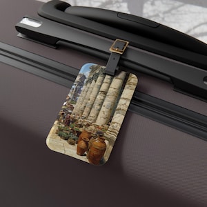 May include: Luggage tag with a painting of Roman columns and pottery. The tag has a black strap and a gold-coloured buckle, attached to a dark grey suitcase. The tag's image shows a classical scene.