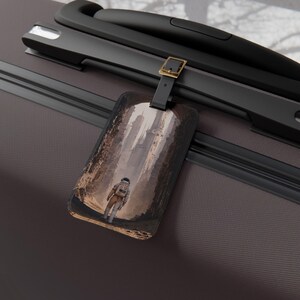 May include: A rectangular luggage tag showing an astronaut walking down a city street with tall buildings. The tag is attached to a black suitcase with a gold buckle.