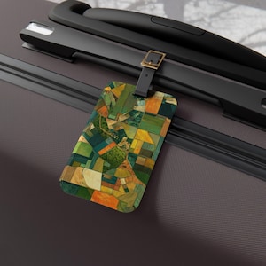 May include: A rectangular luggage tag featuring an abstract design of fields in green, yellow, and brown. The tag is attached to a black strap with a gold buckle, resting on a dark grey suitcase. The design evokes a patchwork of fields.