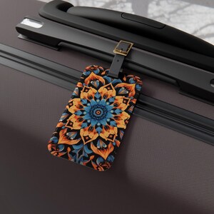 May include: A rectangular luggage tag with a black leather strap and buckle. The tag features a vibrant floral mandala design in shades of blue, orange, and yellow against a black background. The tag is attached to a gray suitcase.