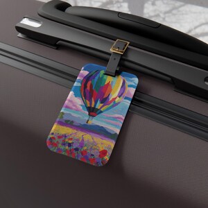Puede incluir: Una etiqueta de equipaje rectangular con un diseño de globo aerostático colorido. El globo está representado en tonos de rojo, amarillo, azul y verde, sobre un fondo de un campo de flores y un cielo azul. La etiqueta está sujeta a una maleta negra.