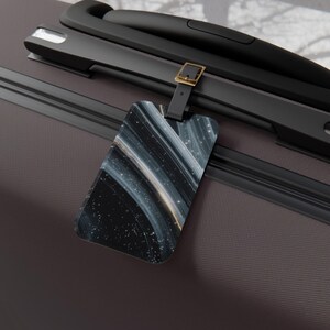 May include: Luggage tag with a black leather strap and gold-colored buckle. The tag features a swirling abstract design in shades of black, white, and gold. The tag is attached to a dark gray suitcase with black handles.