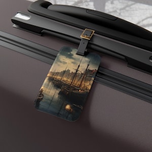 May include: Luggage tag with a scenic harbor illustration. The tag features a rectangular shape with a black strap and a gold-colored buckle. The artwork depicts boats and buildings under a twilight sky, creating a vintage travel aesthetic.