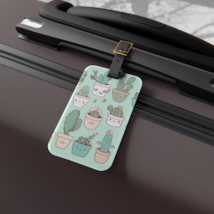 May include: A rectangular luggage tag with a light green background featuring a pattern of potted cacti with smiling faces. The tag is attached to a black suitcase handle with a leather strap and gold buckle.