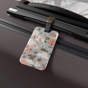 May include: A rectangular luggage tag with a whimsical forest design featuring clouds, trees, and mushrooms in soft pastel colors. The tag is attached to a black suitcase with a leather strap and gold buckle.