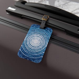 May include: A luggage tag with a blue and white concentric circle design. The tag is rectangular with rounded corners and is attached to a black strap with a gold-colored buckle. The tag is resting on a gray suitcase.