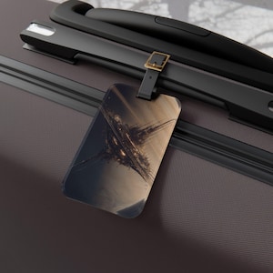 May include: A rectangular luggage tag with a black leather strap and gold buckle. The tag features an abstract design in shades of brown, gold, and black. The tag is attached to a dark gray suitcase with black handles.