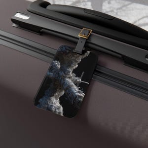 May include: A black luggage tag with a detailed, futuristic architectural design against a black background. The tag is attached to a dark gray suitcase with black handles and a gold buckle.