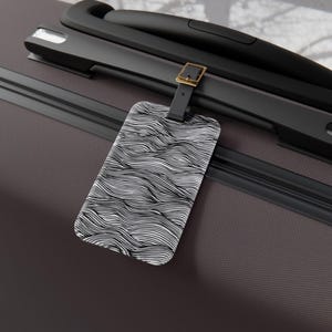 Puede incluir: Una etiqueta de equipaje con una correa de cuero negro y una hebilla dorada. La etiqueta tiene forma rectangular con un patrón ondulado en blanco y negro. La etiqueta está sujeta a una maleta gris oscuro con asas negras.