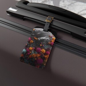 May include: A luggage tag with a black strap and gold buckle, featuring a colorful abstract design. The tag's design includes shades of pink, orange, teal, and yellow against a dark background. The tag is attached to a gray suitcase.