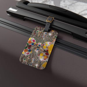 Può includere: Un'etichetta per bagagli con un design astratto e colorato con sfere a pois di vari colori. L'etichetta è rettangolare con angoli arrotondati e un cinturino in pelle nera con fibbia dorata.