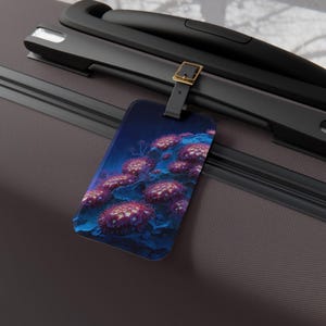 May include: A rectangular luggage tag with an underwater coral reef design. The tag displays pink and purple coral against a dark blue backdrop. It's attached to a black suitcase handle with a leather strap and a gold-coloured buckle.