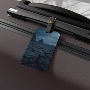May include: Luggage tag featuring a landscape image of cracked earth and rock formations under a cloudy sky. The tag is attached to a black handle with a gold-coloured buckle. The tag is rectangular with rounded corners.