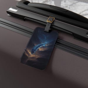 May include: A black luggage tag featuring a blue and white illustration of a centipede-like creature against a dark, cloudy background. The tag is attached to a black suitcase handle with a gold buckle.