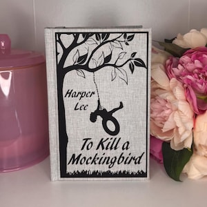 May include: A hardcover book with a light gray linen cover. The cover features a black illustration of a tree with a child swinging from a rope swing, and the title "To Kill a Mockingbird" by Harper Lee.