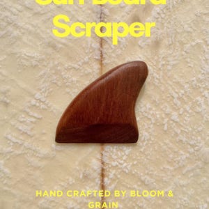May include: A wooden surfboard scraper, curved and brown, with the text "Surf Board Scraper" and "Hand Crafted by Bloom & Grain" in yellow. The scraper is set against a textured, light-coloured background.