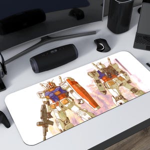 Può includere: Un tappetino per mouse rettangolare bianco con un'illustrazione ad acquerello di due robot Gundam. I robot sono nei toni del bianco, arancione, blu e giallo. Un controller nero, cuffie e altri oggetti tecnologici circondano il tappetino.
