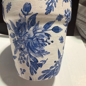 May include: A tapered flower pot featuring a white base with a blue floral pattern. The surface displays a textured finish with detailed illustrations of various blossoms, leaves, and stems wrapping around the exterior of the cylindrical container.