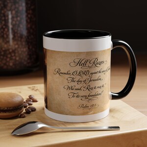 May include: A white ceramic mug with a black interior and handle. The mug features a parchment-style design with the text "Hell Razers" and a quote from Psalms 137:7. A spoon and a pastry are in the foreground.