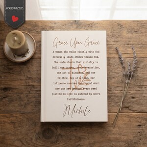 May include: A white hardcover book featuring the text Grace Upon Grace, a religious quote, and the name Michele in black and brown script. The cover includes a cross and heart graphic. A beige candle and dried lavender sprigs are nearby.