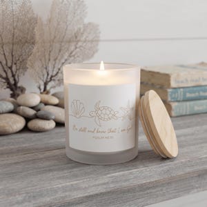 May include: A frosted glass candle with a lit wick and a light wood lid. The label features a line art illustration of a seashell, a sea turtle, and a starfish, with the text: Be still and know that I am God, PSALM 46:10.
