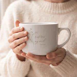 May include: A white ceramic mug featuring a delicate line drawing of wildflowers and the black cursive text "He restores my soul" followed by "PSALM 23:3". The mug has a wide, rounded body and a curved handle on the right side, held in both hands.