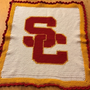 May include: A handmade crochet blanket featuring the letters "SC" in red, with a yellow outline, on a white background. The blanket has a red and yellow scalloped border. The blanket is likely a fan item.
