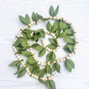 May include: A decorative garland featuring green felt leaves and natural wooden beads. The leaves are arranged along the garland, creating a natural, organic look. This garland is suitable for home decor and seasonal displays.