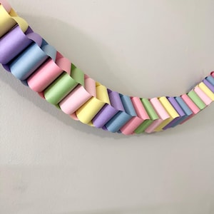 May include: A pastel-colored paper chain garland in shades of pink, yellow, green, blue, and purple. The chain is made of looped paper strips, creating a decorative, woven pattern. The garland is against a plain white wall.