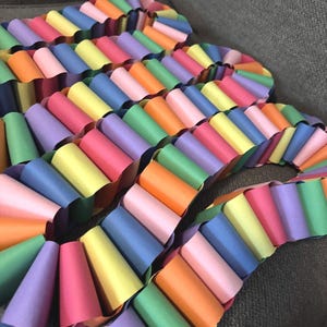 May include: A colorful paper garland featuring a repeating pattern of rolled paper cones in various colors. The garland includes shades of pink, orange, yellow, green, blue, and purple. The decorative item is arranged in a wavy pattern.