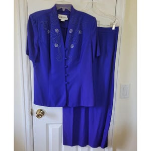 Puede incluir: Un traje de dos piezas azul cobalto. La chaqueta de manga corta tiene una parte delantera abotonada y diseños florales y de remolino bordados. La falda a juego es de estilo recto, hasta la rodilla. El traje está colgado en una percha.