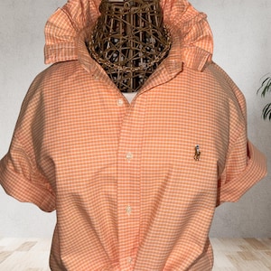 May include: An orange and white plaid button-down shirt with short sleeves, knotted at the waist. The shirt features a small embroidered logo and the text "Relaxed Fit: L/XL".