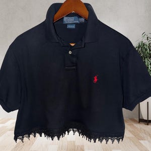 May include: A black short-sleeved polo shirt featuring a collar, a two-button placket, and a red embroidered logo on the left chest. The hem is finished with a decorative black lace trim. The interior neck label reads Polo by Ralph Lauren.