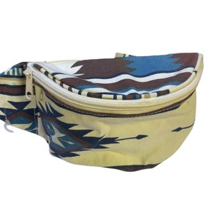 May include: A waist bag featuring a cream-colored base with a geometric pattern in shades of dark brown, navy blue, and white. The bag has a curved top zipper closure with a light-colored zipper tape and a matching pull tab on the left side.