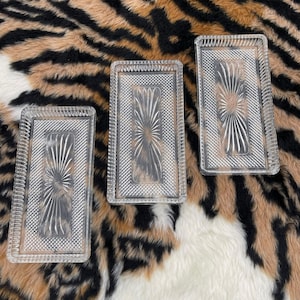 May include: Three rectangular clear glass trays with raised edges. Each tray features a central sunburst pattern surrounded by a textured cross-hatch border and a ribbed outer rim. The glass is transparent and reflective, showcasing a geometric design.