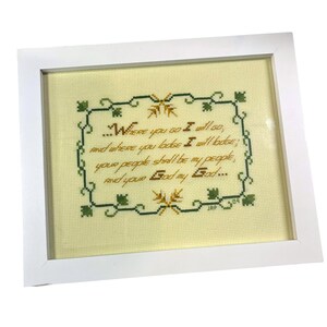 May include: Framed cross-stitch artwork with a white frame. The artwork features a quote in brown thread on a light yellow background, surrounded by a green and brown decorative border. The text reads, "Where you go I will go..." and is signed "JAP 09".