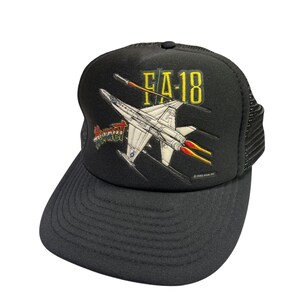 May include: A black trucker hat featuring a graphic print of an F/A-18 Hornet fighter jet in flight with yellow exhaust flames. The text FA-18 is printed in yellow above the jet, and the word Hornet is printed in red, white, and blue to the left.