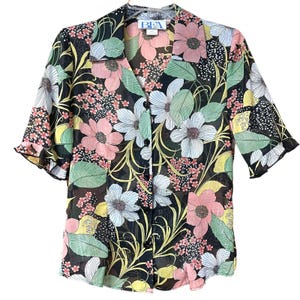 May include: A black button-down blouse with a floral pattern. The blouse features large pink, white, and blue flowers, green leaves, and yellow vines. The sleeves are short and ruffled. The label reads "BEA".