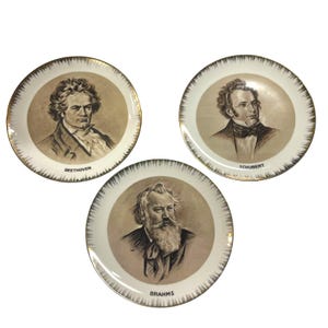 May include: Three round ceramic decorative plates featuring sepia-toned portraits of classical composers. Each plate has a gold-trimmed edge with a radiating spike pattern. The plates are labeled with the names BEETHOVEN, SCHUBERT, and BRAHMS.