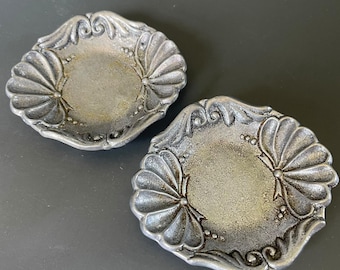 May include: Two small, round, silver-toned metal dishes featuring a textured, weathered finish. Each dish has a raised, scalloped border with embossed shell-like patterns and small bead accents along the edges, creating a decorative, circular rim.
