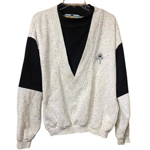 May include: A vintage sweatshirt with a black mock turtleneck and a gray speckled V-neck. The sleeves have black panels. A small embroidered design is on the chest. The sweatshirt is hanging on a black hanger.