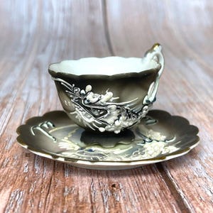 May include: A small porcelain teacup and saucer set featuring a dark grey and white gradient finish. The exterior of the cup and the surface of the saucer are decorated with raised, three-dimensional white floral and vine relief patterns.