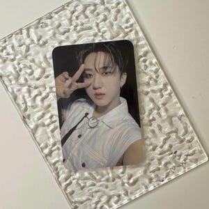 May include: A photograph of a person in a white sleeveless shirt, making a peace sign with their fingers. The photo is in a clear, textured glass frame. The person has dark hair and is looking at the camera.