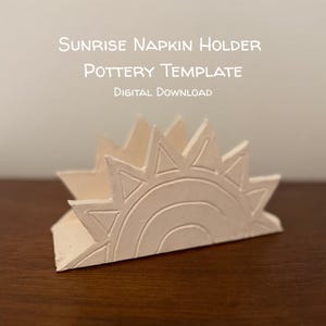 May include: A cream-colored pottery napkin holder in a sunrise design. The holder features a sun with rays and a rainbow, with the text "Sunrise Napkin Holder Pottery Template Digital Download". The holder is on a wooden surface.