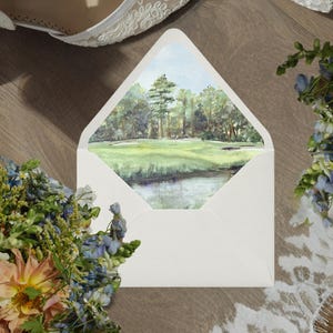 以下が含まれることがあります： ゴルフコースの風景を描いた水彩画が入った白い封筒。封筒は、フラワーアレンジメントと白いレースの靴のペアに囲まれています。シーンには、緑のフィールド、木々、水域が含まれています。
