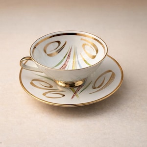 May include: A white teacup and saucer set with gold trim and decorative gold swirls. The cup and saucer have a delicate design with colorful accents. The set is placed on a neutral background.