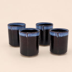 May include: A set of four cylindrical ceramic cups featuring a dark brown, textured grid pattern on the lower body and a smooth, contrasting light blue glaze around the upper rim. Each cup has a vertical seam detail along the side.