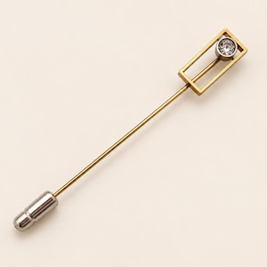 May include: A gold-toned lapel pin featuring a long, slender needle with a silver-toned cylindrical end cap. The head of the pin consists of a rectangular gold frame containing a single, small, clear, round faceted gemstone set in silver.